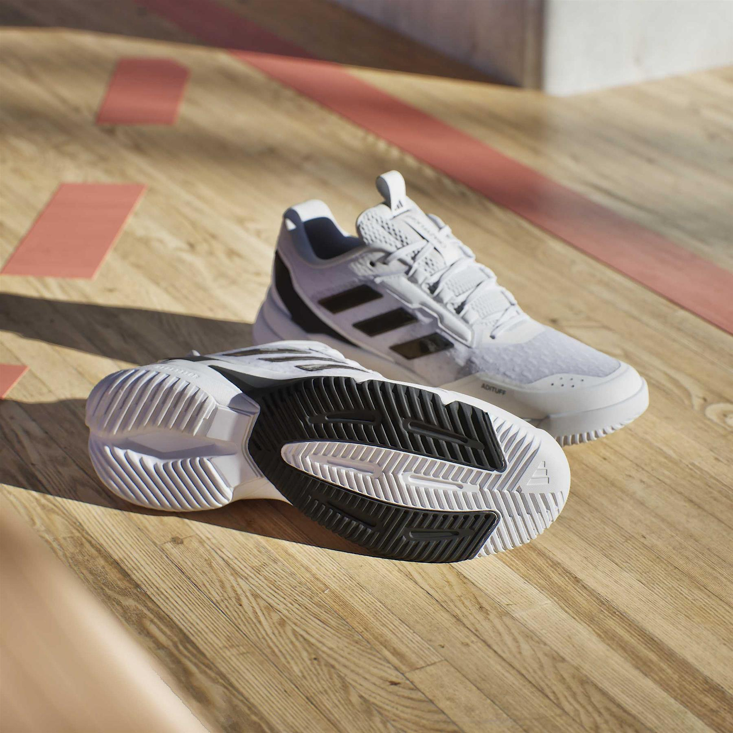 A pair of adidas Crazyflight 6 Men's Indoor Badminton Shoes in white are displayed on a wooden court floor—one upright, the other showing its sole—with sunlight casting gentle shadows.