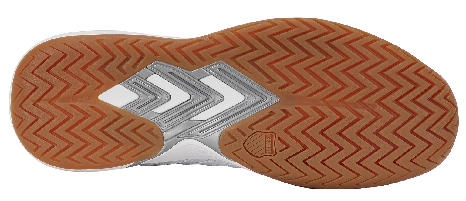The image shows the bottom of the K-Swiss Ultrashot Light Indoor Men's Badminton Shoe in White, Black, and Silver. It features a brown non-marking gum rubber outsole with a herringbone tread pattern, silver arrow-like designs on the midfoot, and a small K-Swiss logo near the arch.