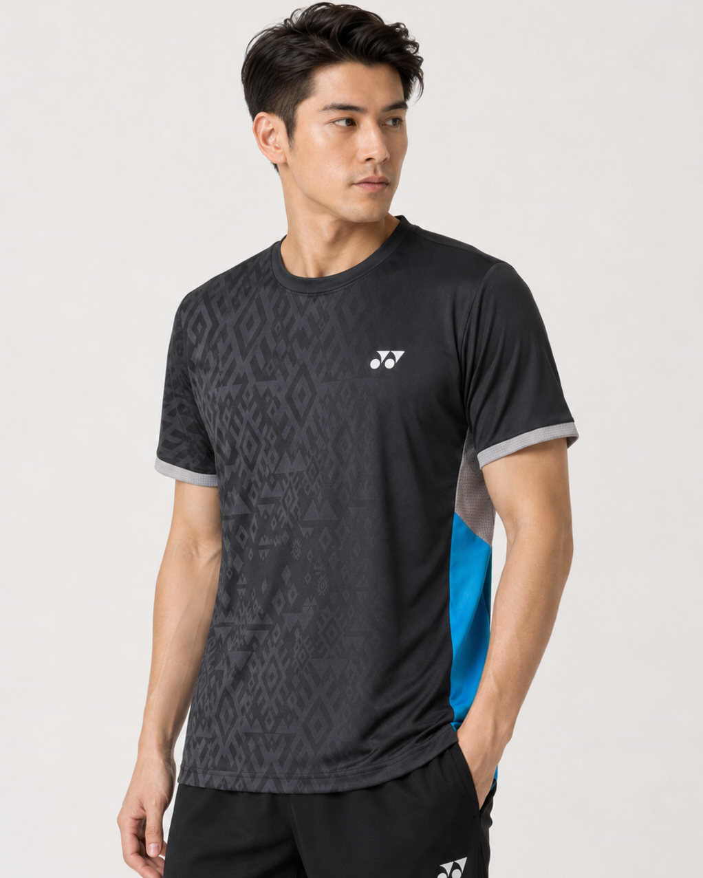 A man wearing the Yonex YTM11 Men's Badminton T-Shirt in black by Yonex, featuring gray trim and blue side panels in Air-Release fabric, stands with one hand in his pocket, looking left against a plain light background.