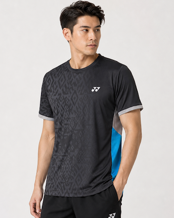 A man wearing the Yonex YTM11 Men's Badminton T-Shirt in black by Yonex, featuring gray trim and blue side panels in Air-Release fabric, stands with one hand in his pocket, looking left against a plain light background.