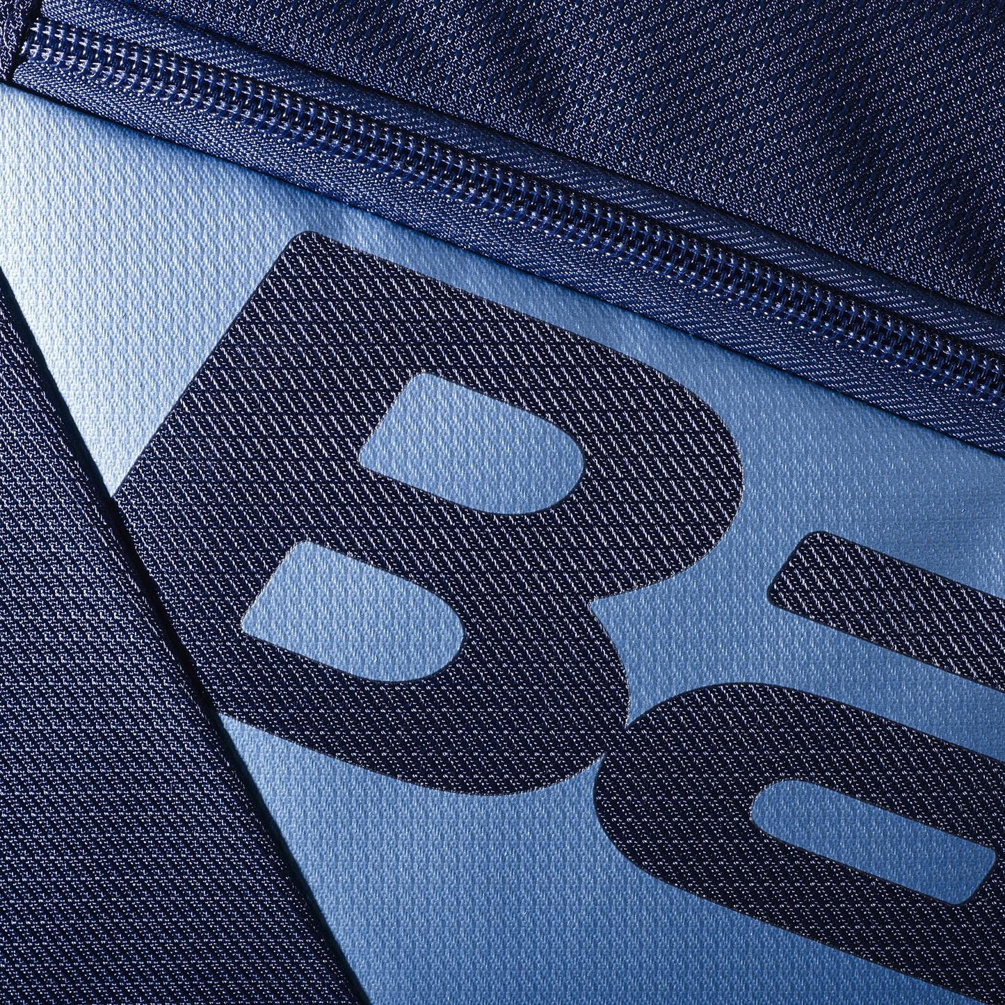 Close-up of a navy blue fabric with a zipper partially visible, mimicking the Babolat RH12 Pure Drive Gen11 12 Racket Bag. The textured material shows partial "Ba" letters in black on the lighter blue section, reflecting the sleek style of this Babolat badminton bag.