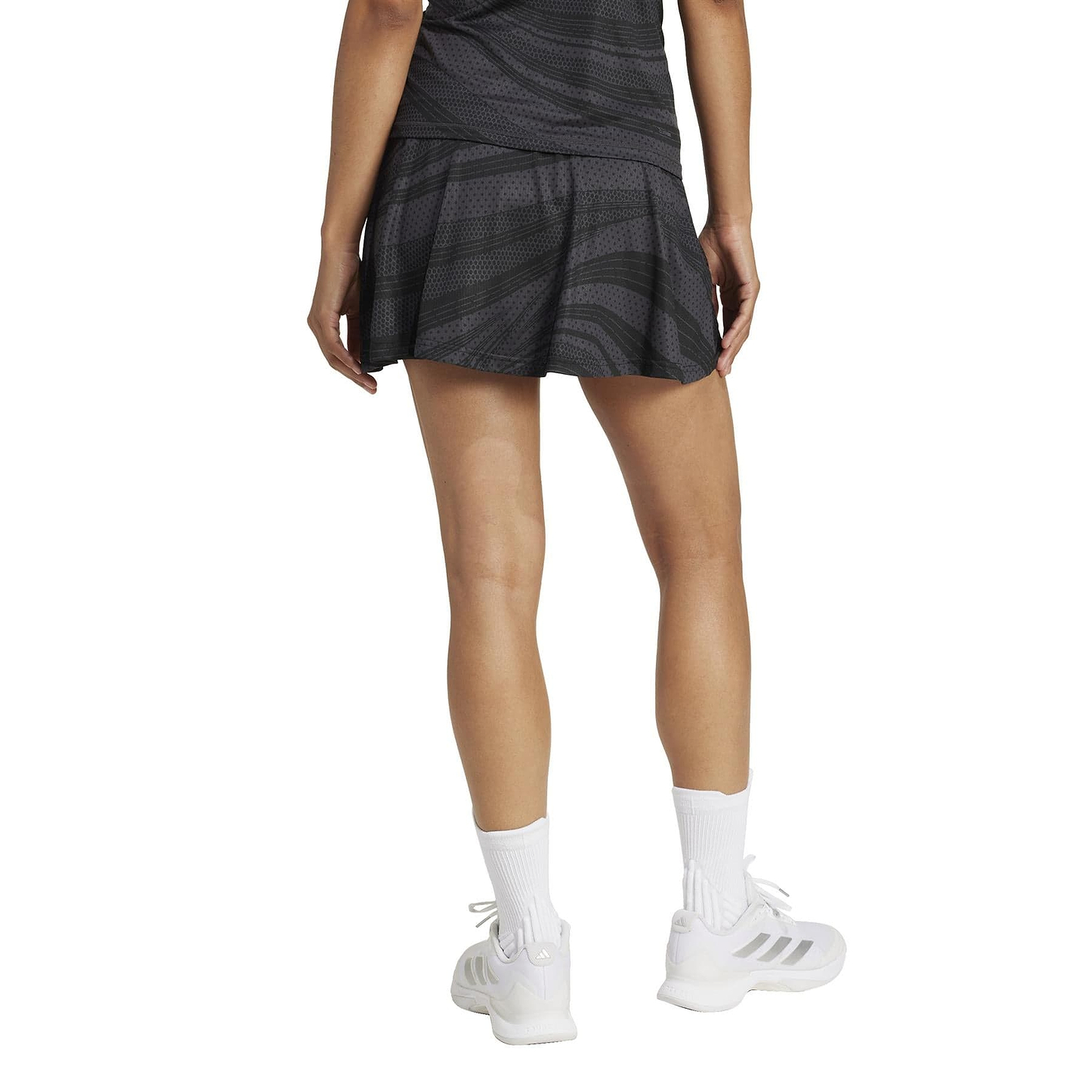 A person in an ADIDAS Women's Club Graphic Badminton Skirt - Black, with white socks and CLIMACOOL sneakers, stands on a light background. The image shows the back view of this stylish recycled-material outfit.