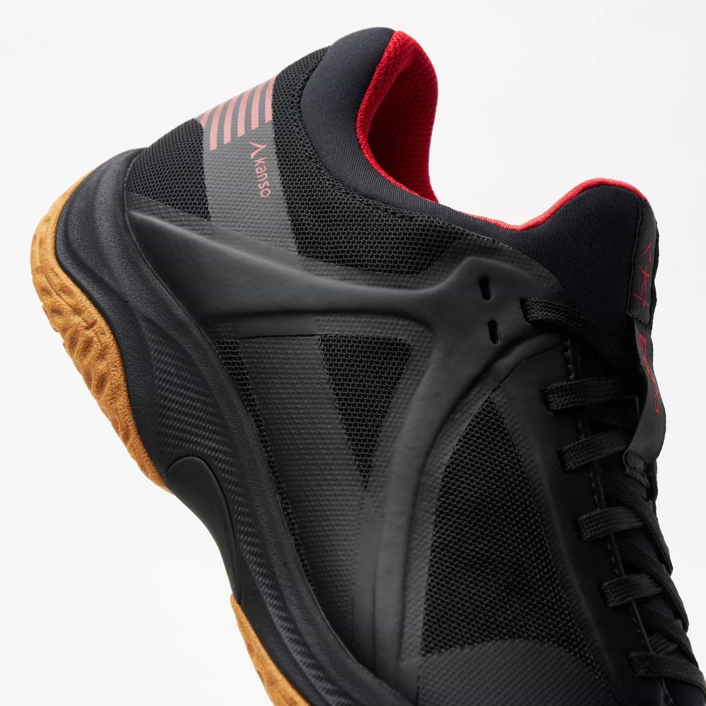Detailed view of the Kanso Equilibrium EQx3 Unisex Badminton Shoes in black, featuring red accents and a brown sole. Made with breathable mesh and Speedlight foam midsole for comfort, it showcases a modern design with stripes and branding on the heel.