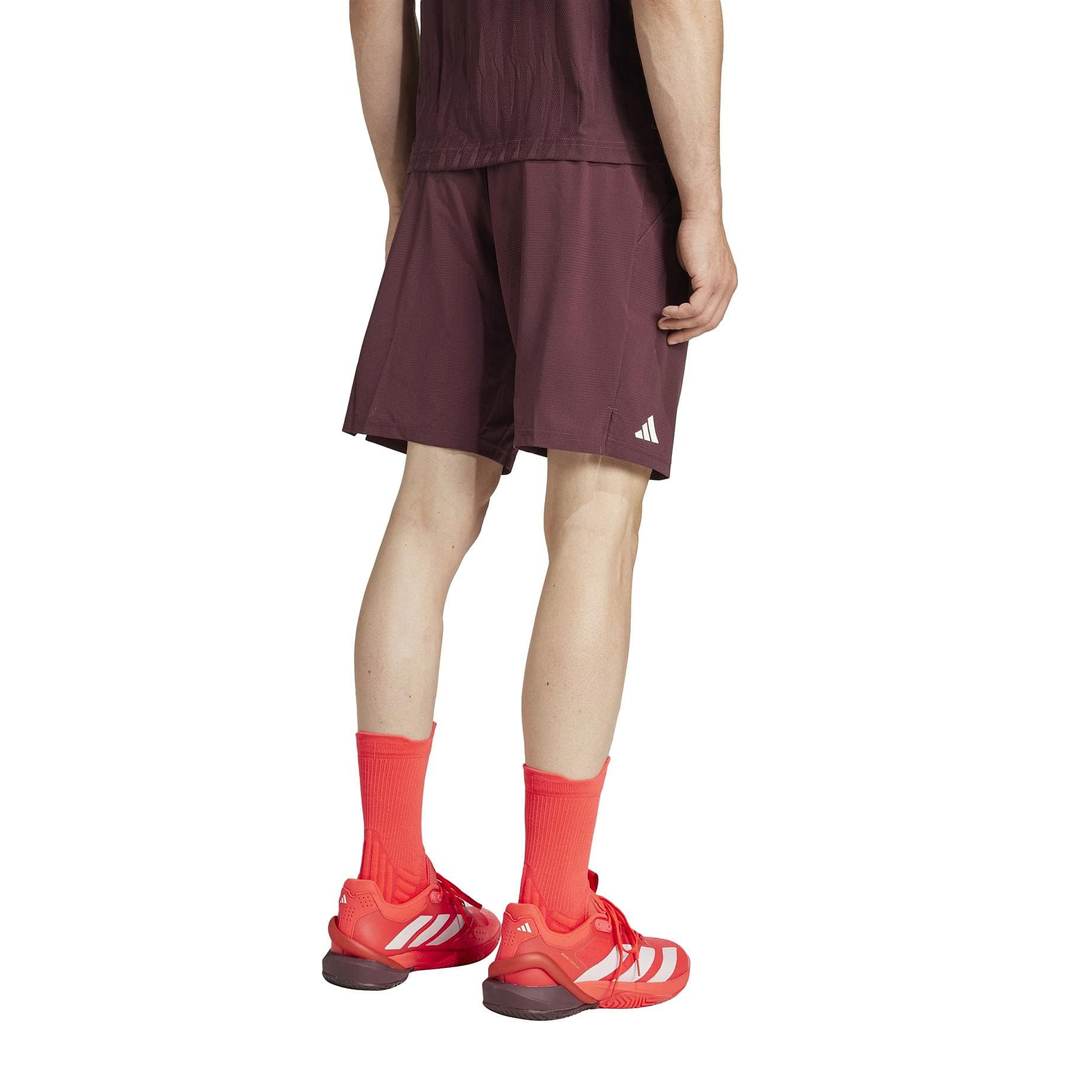 The individual faces away from the camera, wearing adidas Ergo Men's Badminton Shorts in ruby, paired with a maroon athletic outfit, bright red socks, and HEAT.RDY sneakers featuring white stripes. This eco-friendly ensemble is showcased against a plain white background.