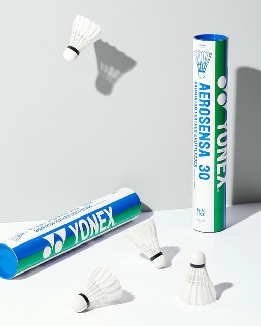 Yonex Aerosensa AS30 Feather Shuttles (Set of 12) by Yonex are shown on a light surface—two shuttlecocks appear mid-air, highlighting flight stability, with two branded tubes displayed, one upright and one lying down.