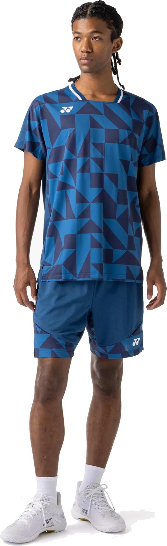 A man wearing a Yonex 10712YX Crew Neck Men's Badminton T-Shirt in Indigo Blue with matching shorts, white socks, and sneakers stands slightly turned to the side against a white background.