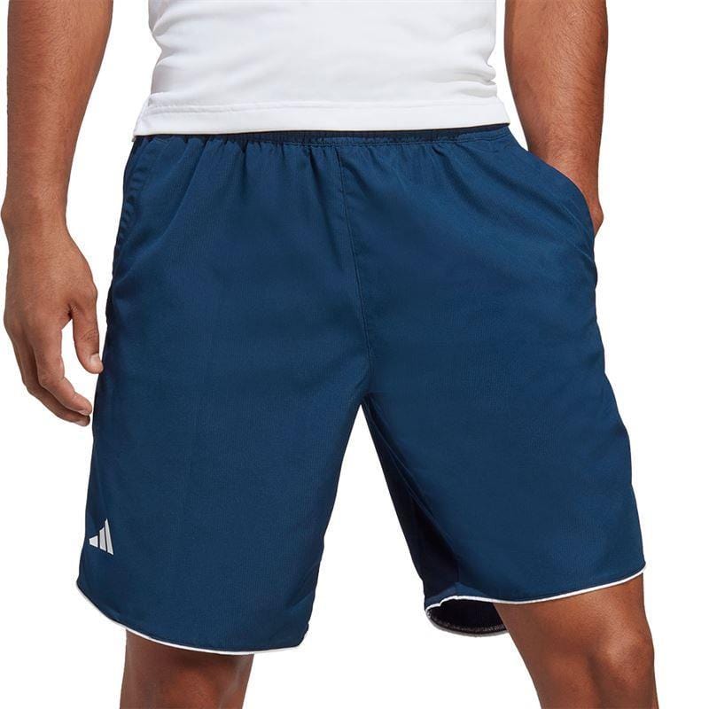 Wearing a white T-shirt, the individual sports the ADIDAS Men's Club 7" Badminton Shorts in navy blue, accented with white trim and adorned with a small logo on the left leg. With hands tucked in pockets against a plain backdrop, they highlight these stylish shorts equipped with AEROREADY technology by adidas.