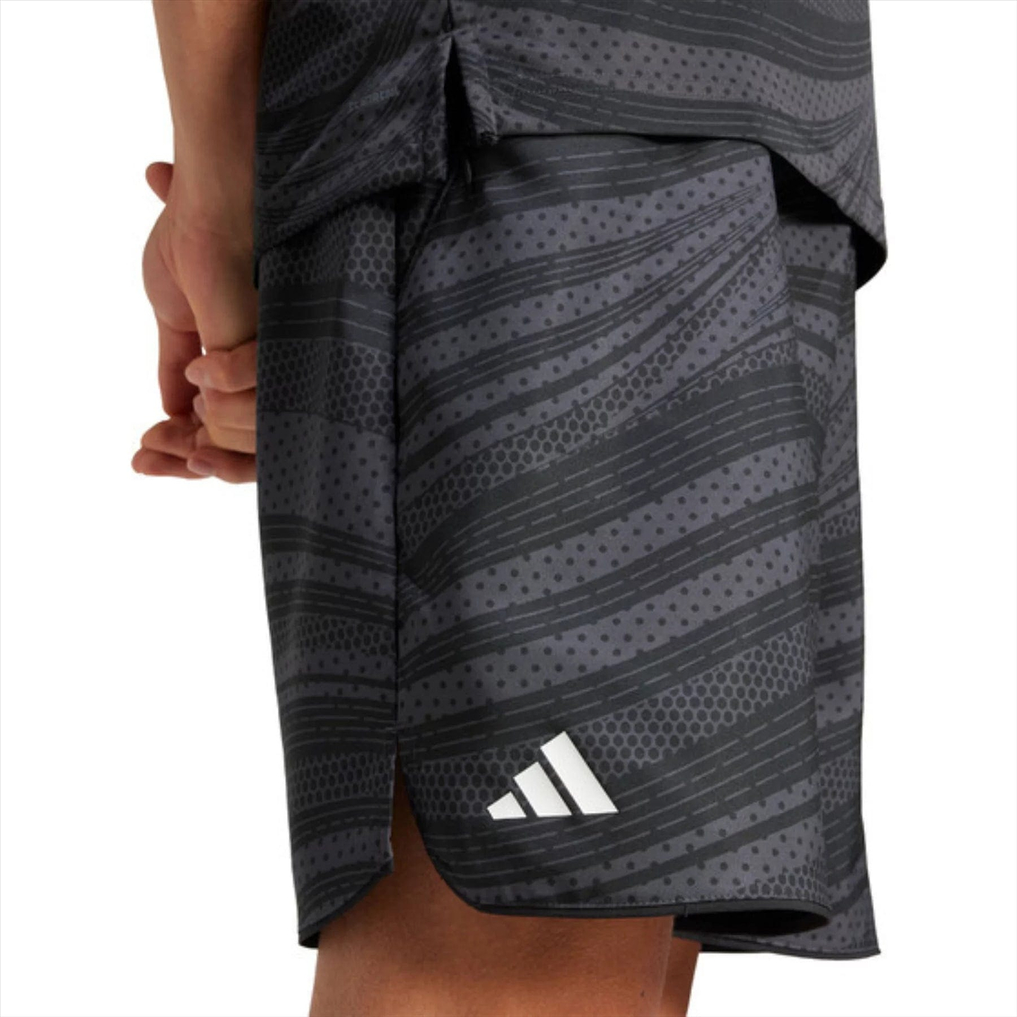 A person in the ADIDAS Club Graphic Men's Badminton Shorts - Black, featuring a white logo on the lower left, wears a dark athletic outfit with gray stripes made from recycled materials and stands with their hands clasped behind their back.