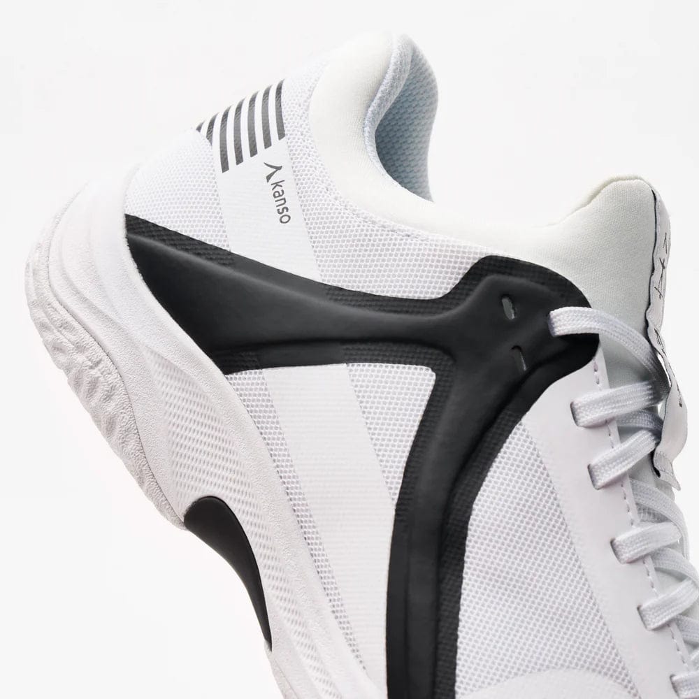 Close-up of the Kanso Equilibrium EQx3 unisex badminton shoe in white/black. It features a textured sole with shock absorption, branded "Kanso" on the side, Speedlight Foam for performance, and parallel black stripes near the top.