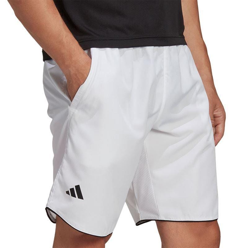 A person wearing ADIDAS Men's Club 7" Badminton Shorts in white, featuring a black logo on the left leg, stands with hands in pockets. The AEROREADY shorts have black trim and are paired with a sleek black top, ideal for an energetic badminton session.