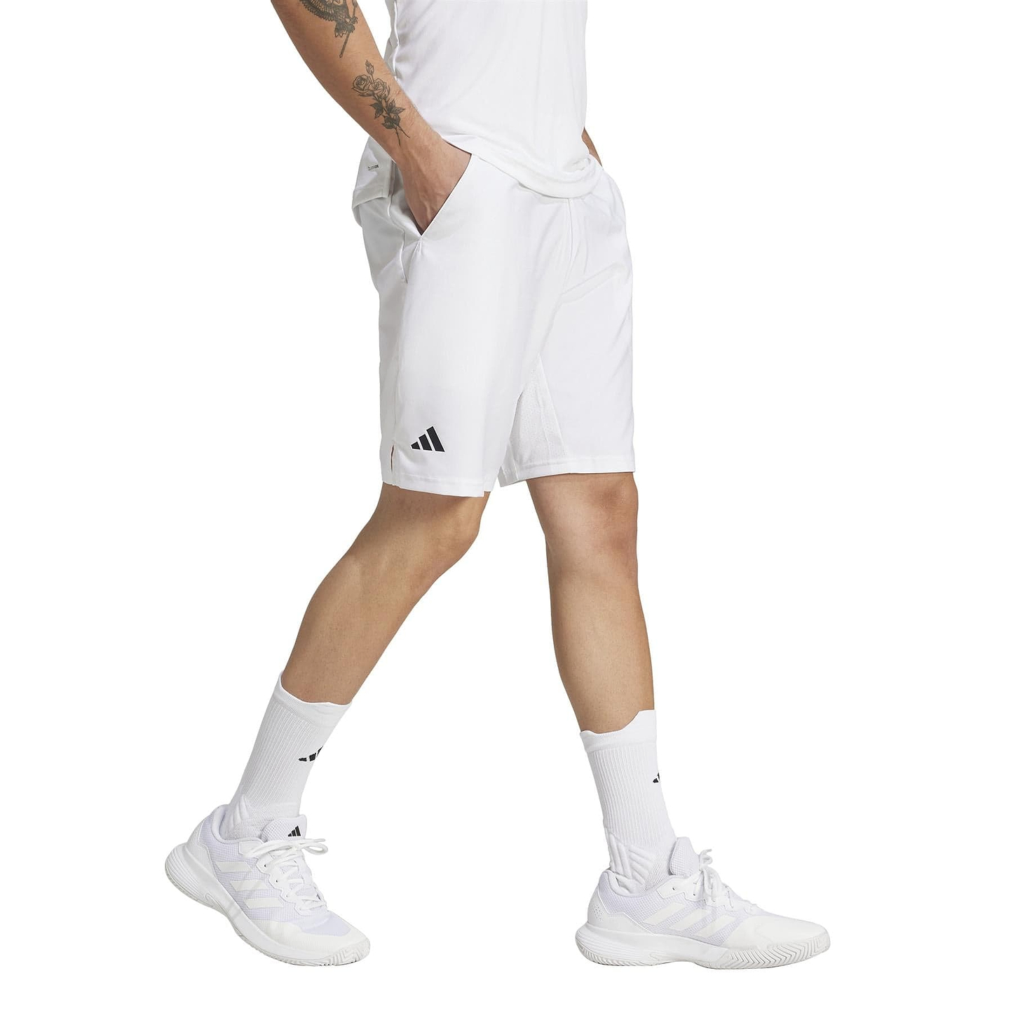 A tattooed individual in white ADIDAS Men's 3 Stripe Badminton Shorts with a black logo and sneakers is shown from the shoulders down. The moisture-wicking fabric ensures comfort as they walk against a plain white background.