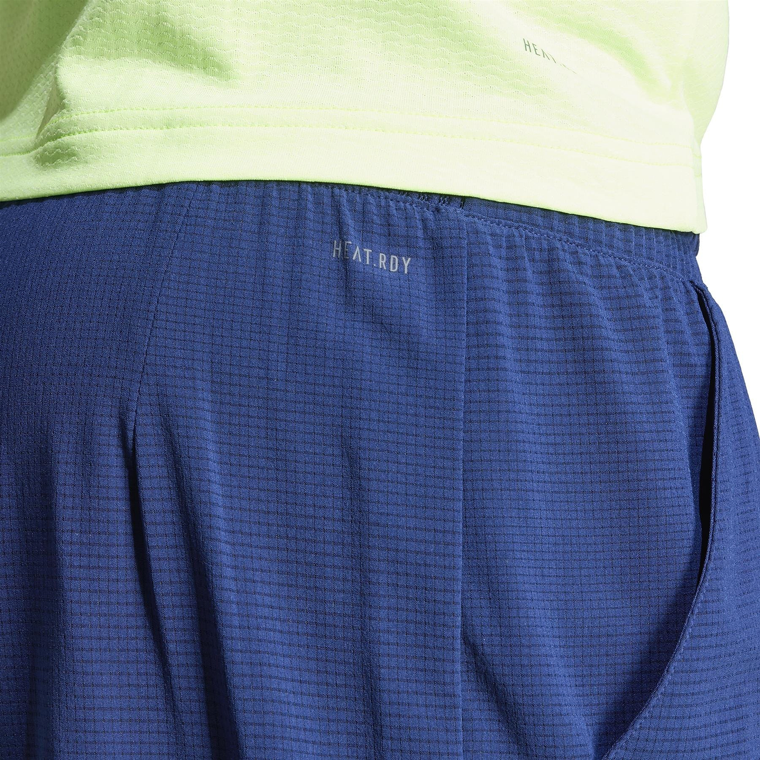 Close-up of a person's lower back wearing a neon green shirt and lightweight navy blue textured shorts. These adidas Ergo Men's Badminton Shorts feature a discreet zipper pocket and "HEAT.RDY" logo, crafted with recycled materials for comfort and sustainability.