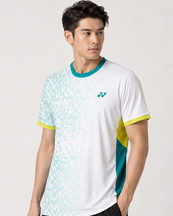 A man with short dark hair wears the Yonex YTM11 Mens Badminton T-Shirt - White by Yonex, which has light green geometric patterns, teal and yellow accents, Air-Release fabric, and a small chest logo, standing against a plain light background.