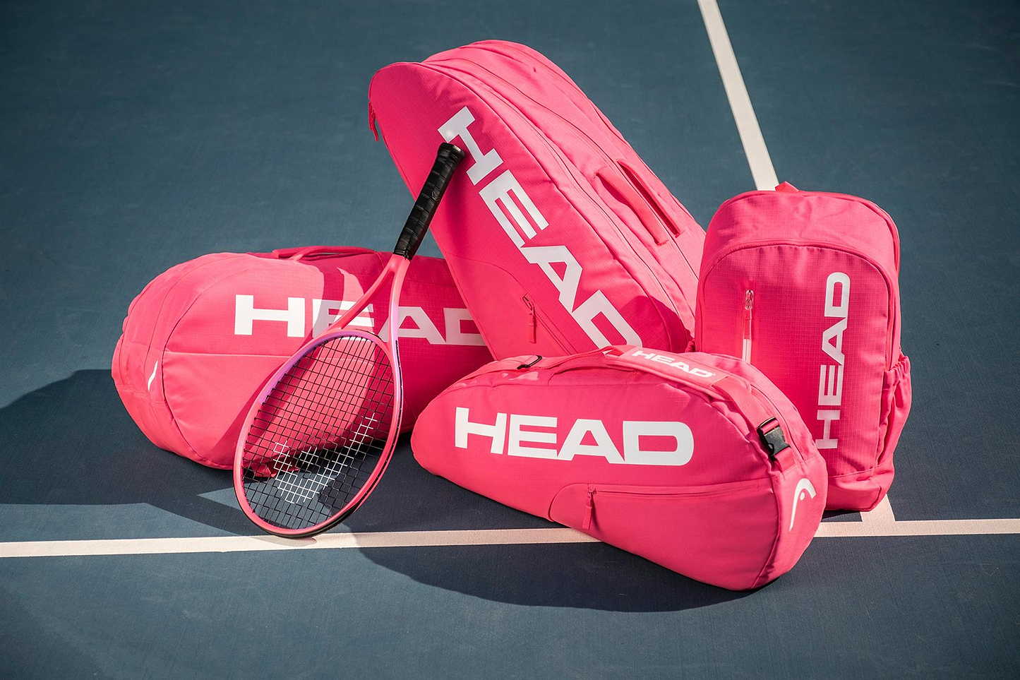 Four HEAD Base 6 Racket Badminton Bag M - Pink by HEAD are arranged on a tennis court—two upright, two lying down—beside a matching pink racket and a white court line. Ideal for badminton or tennis gear.