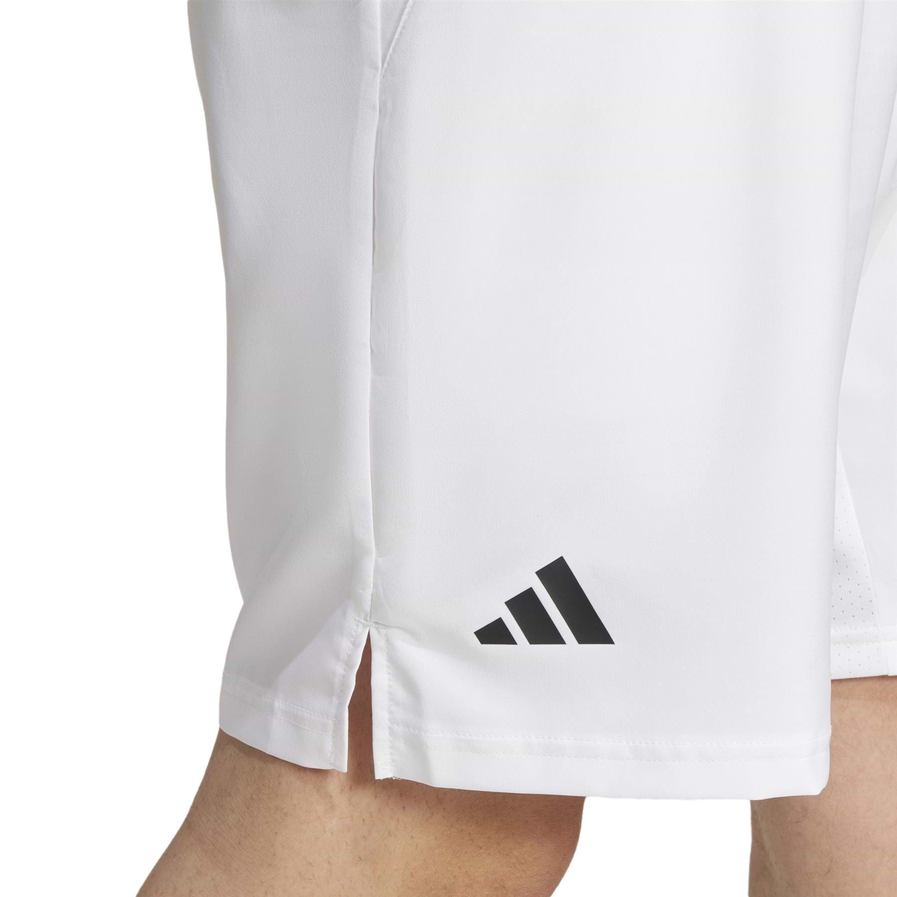Close-up of someone wearing ADIDAS Men's 3 Stripe Badminton Shorts in white, showcasing the black logo near the hem. These shorts have moisture-wicking properties and a small side slit for cool comfort on the court, focusing on the lower part of the shorts and legs.