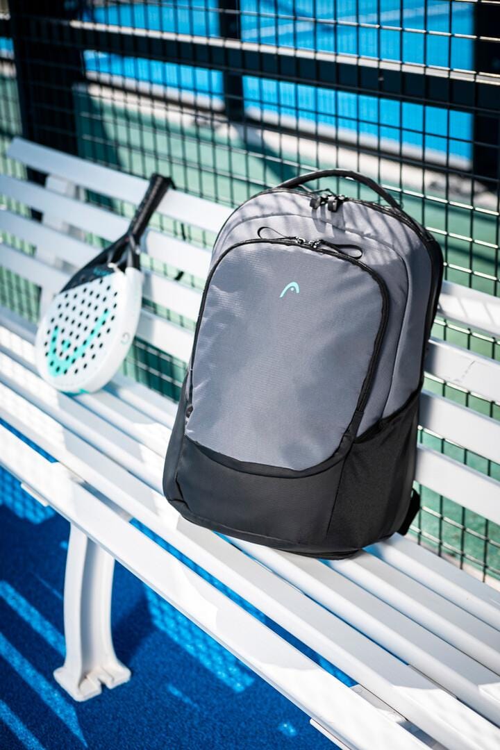 A HEAD Pro X Badminton Backpack 30L in Black/Dark Grey rests on a white bench beside a padel racket with a white face and black handle. The bench is near a black wire fence on the blue court, and the eco-friendly setup, made from recycled PET bottles, casts shadows in bright daylight.