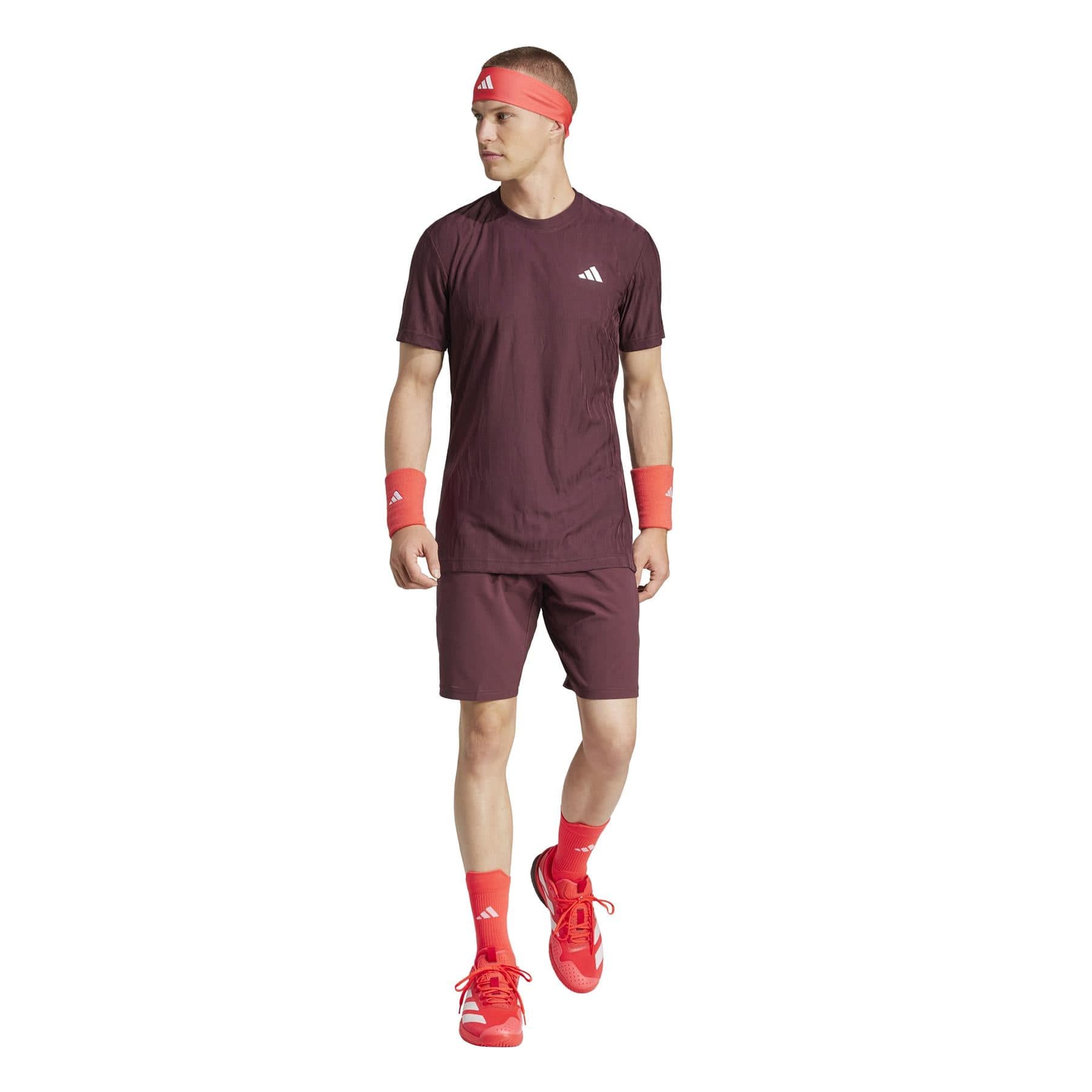 On a white background, a person confidently wears adidas Ergo Men's Badminton Shorts in Ruby, complemented by a maroon athletic set including a matching T-shirt, red sneakers, headband, wristbands, and socks while gazing slightly to the side.