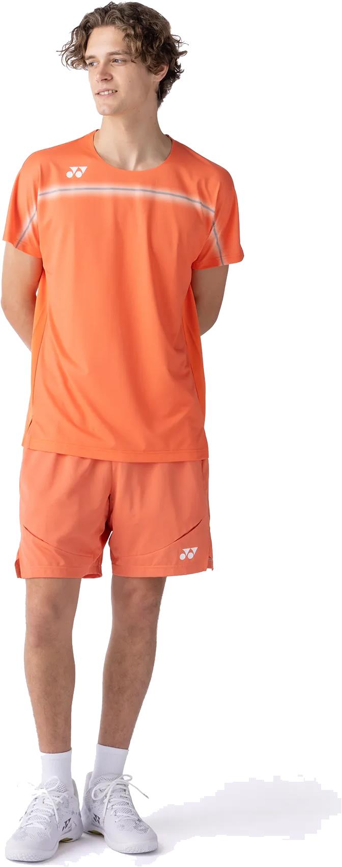A young man stands with his hands behind his back, wearing the Yonex 10711YX Crew Neck Men's Badminton T-Shirt in Cyber Orange—showcasing Yonex's stylish performance apparel against a white background.