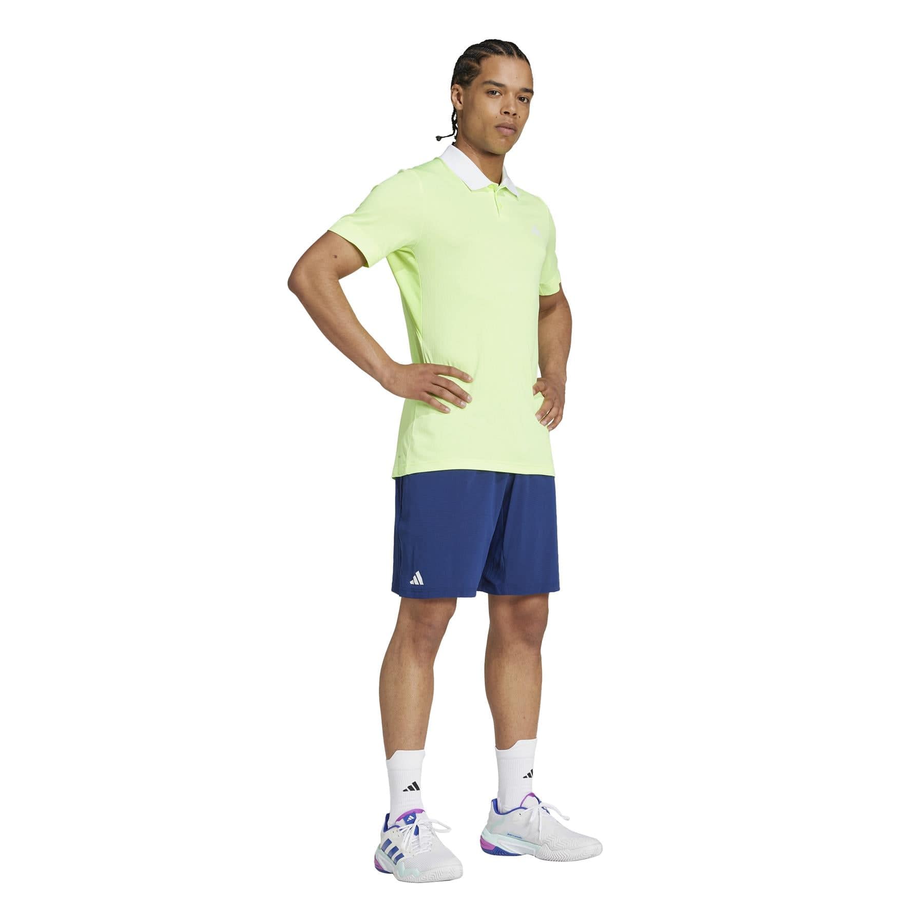 On a plain white background, a person with braided hair poses, hands on hips. They're wearing a light green polo shirt paired with adidas Ergo Men's Badminton Shorts in navy, crafted from lightweight recycled materials. White sneakers complete the ensemble.