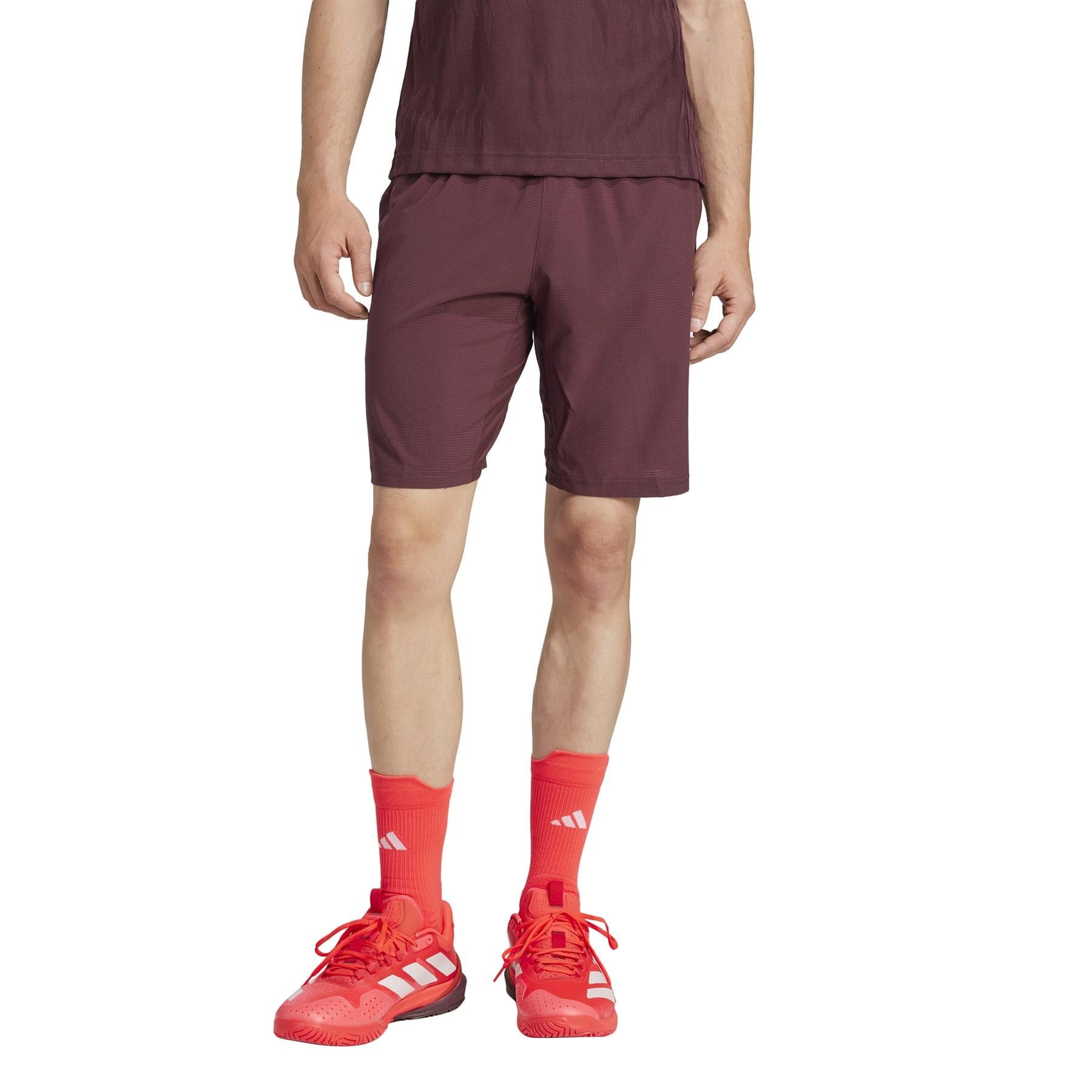 A person in a burgundy top and matching ADIDAS Ergo Men's Badminton Shorts - Ruby, crafted from recycled materials, bright red-orange socks, and red athletic shoes stands against a white background; only their lower half is visible.