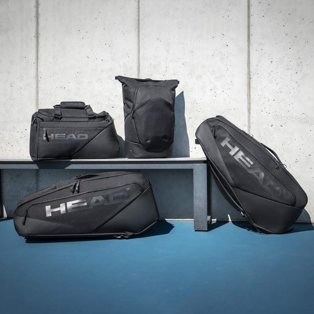 Four black HEAD sports bags, including the HEAD Pro X 12 Racket Badminton Racket Bag XL - Black, of various shapes and sizes are arranged on and in front of a bench against a concrete wall and blue floor, shown in natural light.