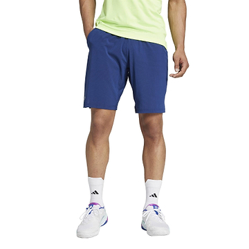 A person wearing a light green shirt and ADIDAS Ergo Men's Badminton Shorts in navy stands against a plain background. They sport white HEAT.RDY socks and sneakers with purple accents, with their hands resting casually in their pockets.