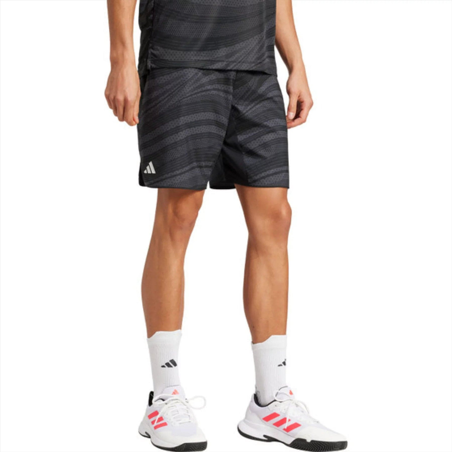 A person is wearing ADIDAS Club Graphic Men's Badminton Shorts in black, coupled with a matching shirt, logo-adorned white socks, and pink-accented white sneakers. The focus is on their athletic attire made from recycled materials against a plain backdrop.