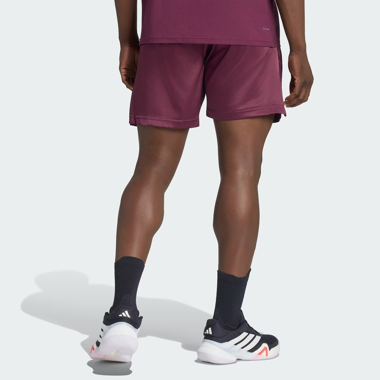 A person stands facing away, wearing adidas Climacool Ergo Pro Men’s Badminton Shorts in maroon, a matching shirt, black socks, and black adidas sneakers with white soles.