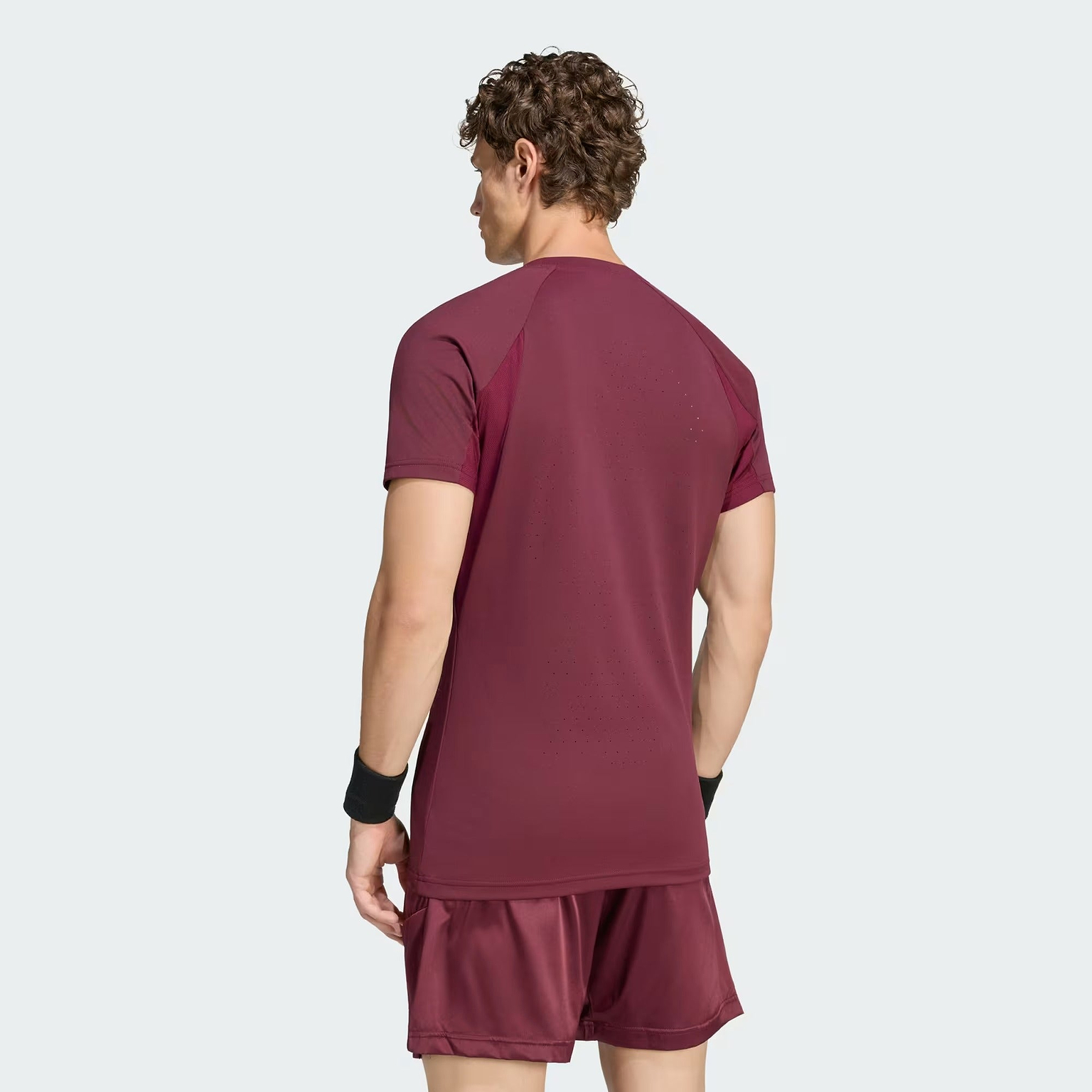 A man with curly hair, wearing a maroon adidas Climacool+ Freelift Pro Men’s Badminton T-Shirt and matching shorts, stands facing away from the camera against a plain light background.
