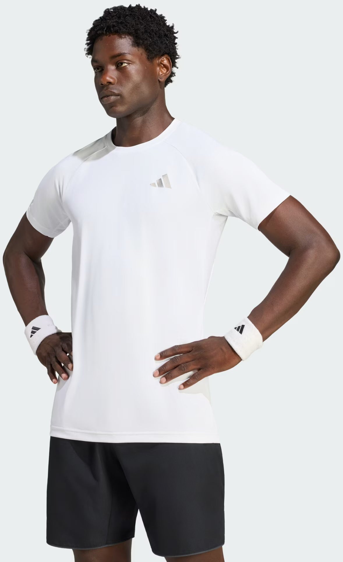 A man stands with hands on hips, wearing an adidas Climacool+ Freelift Pro Men’s Badminton T-Shirt in white—an advanced, quick-drying sports tee—paired with black shorts and white adidas wristbands, looking serious.