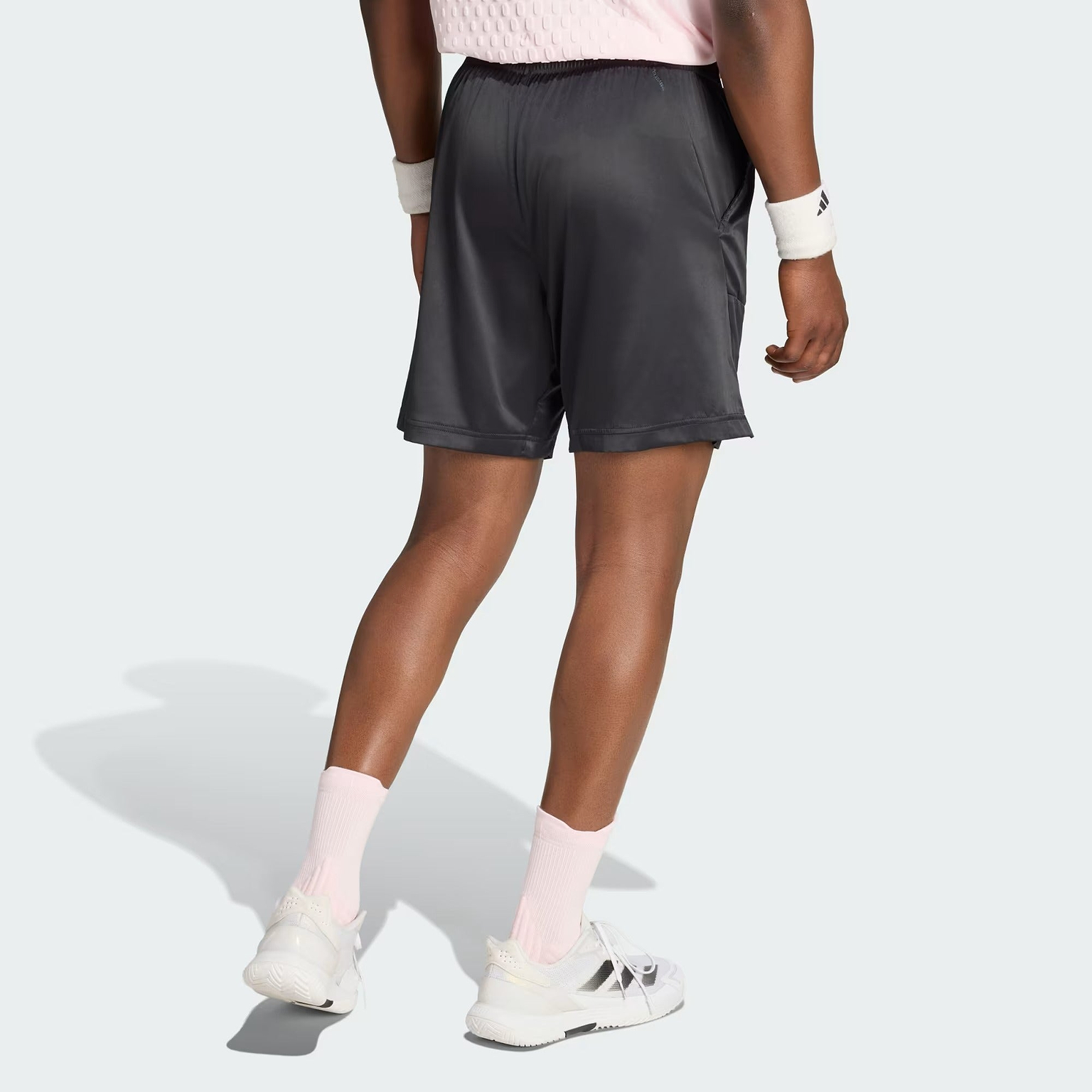 A person wearing a light pink shirt, adidas Climacool Ergo Pro Mens Badminton Shorts in black with an ergonomic cut, light pink socks, white sneakers, and white wristbands stands facing away, casting a shadow on a light background. The outfit’s Climacool fabric from adidas ensures lasting comfort during activity.