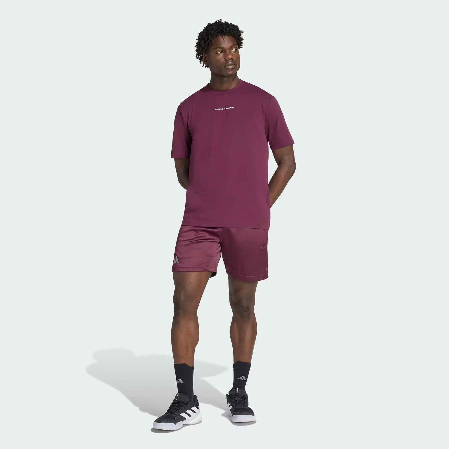 A man stands against a light background wearing a matching maroon adidas t-shirt and ADIDAS Climacool Ergo Pro Men’s Badminton Shorts in maroon. He pairs these with black socks and black-and-white adidas sneakers, has his hands behind his back, and looks slightly to the side.