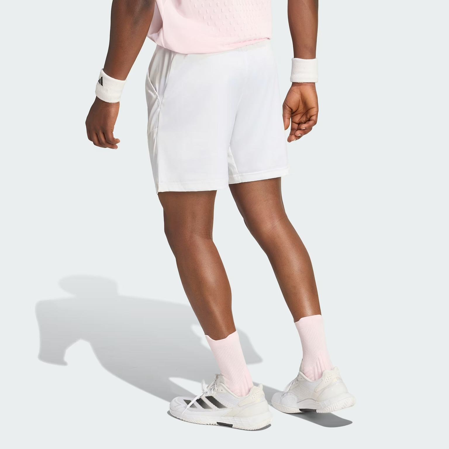 A person wearing a light pink shirt, white adidas Climacool Ergo Pro Men’s Badminton Shorts, pink socks, white sneakers, and wristbands stands on a light background, facing away with their arms relaxed at their sides.