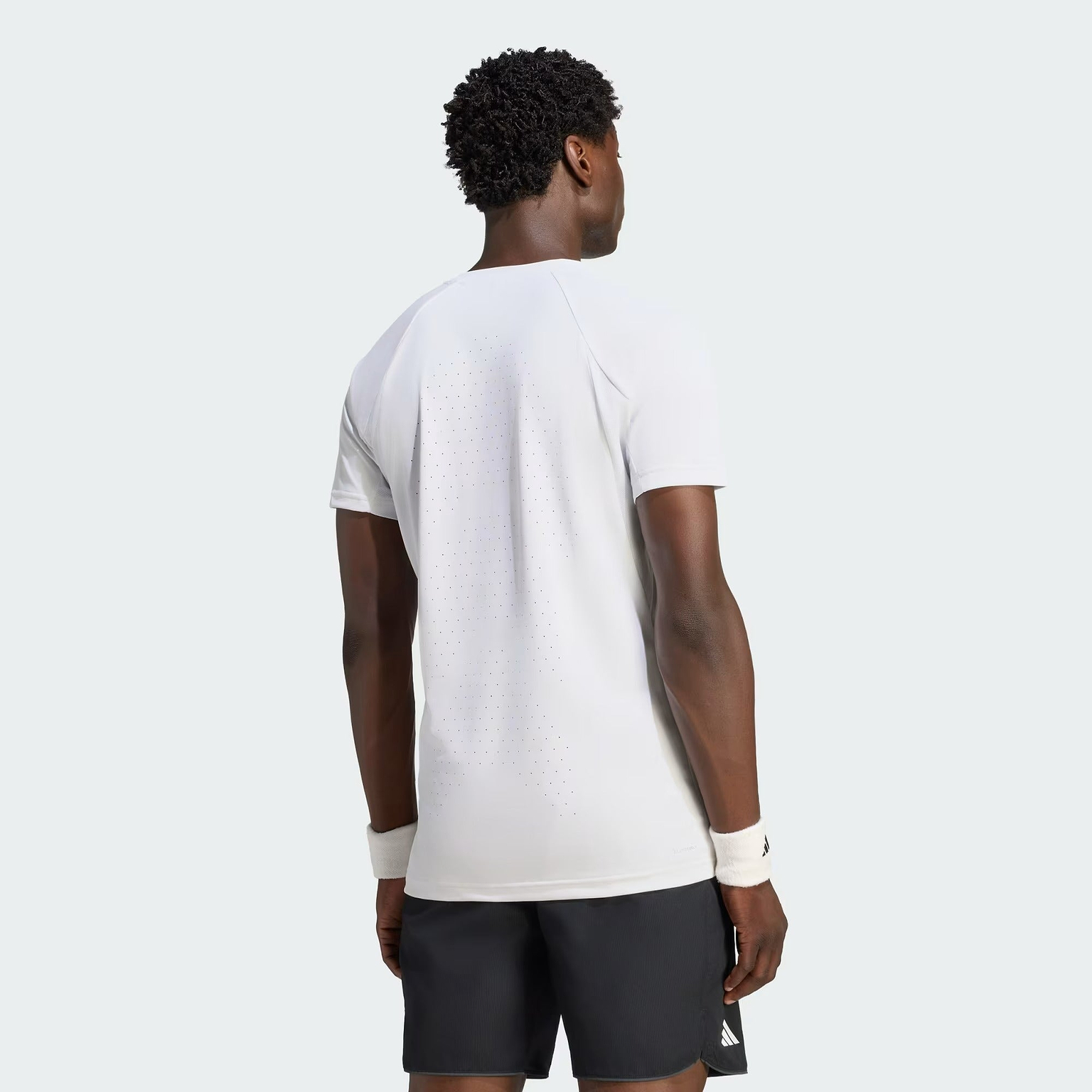 A man stands facing away, wearing the adidas Climacool+ Freelift Pro Men’s Badminton T-Shirt in white—a high-performance, quick-drying sports tee—paired with black shorts and white wristbands against a light gray background.