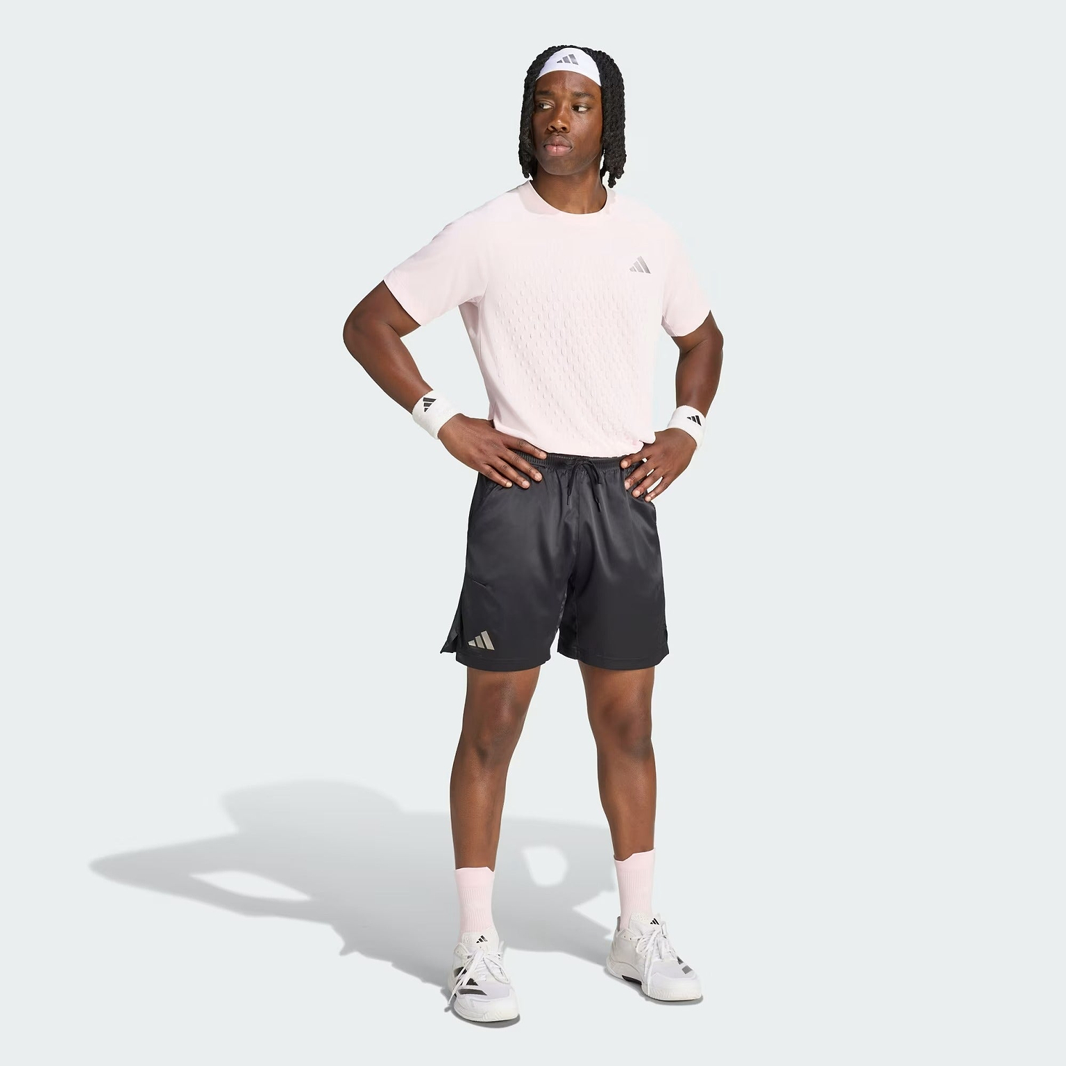 A person stands with hands on hips, wearing a light pink Adidas shirt, black ADIDAS Climacool Ergo Pro Men’s Badminton Shorts featuring ergonomic construction, white sneakers, white socks, wristbands, and a white Adidas headband against a light gray background.