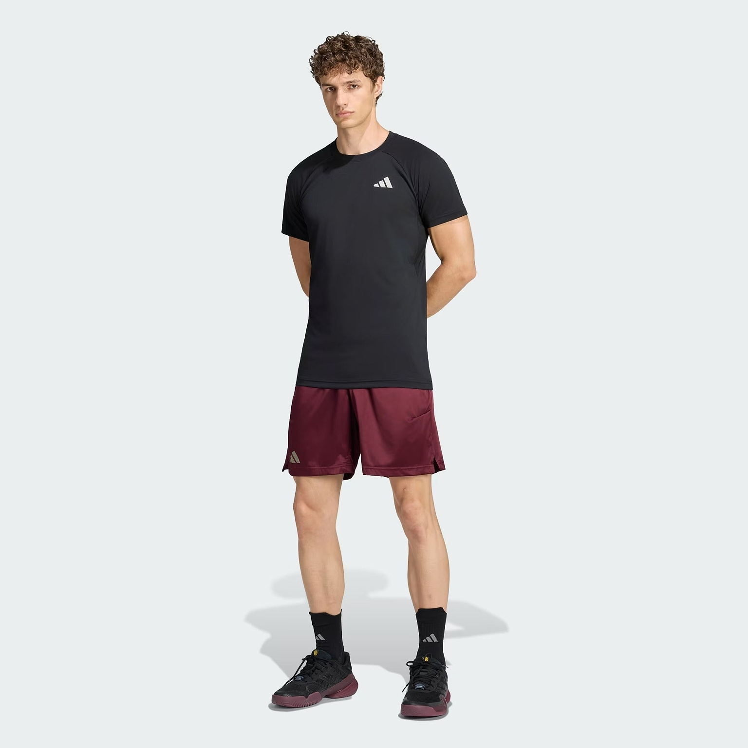 A man stands against a light background wearing an adidas Climacool+ Freelift Pro Men’s Badminton T-Shirt in black, paired with maroon shorts, black socks, and black shoes—all featuring adidas logos. The performance badminton shirt is engineered with advanced cooling technology to keep you comfortable during intense matches.