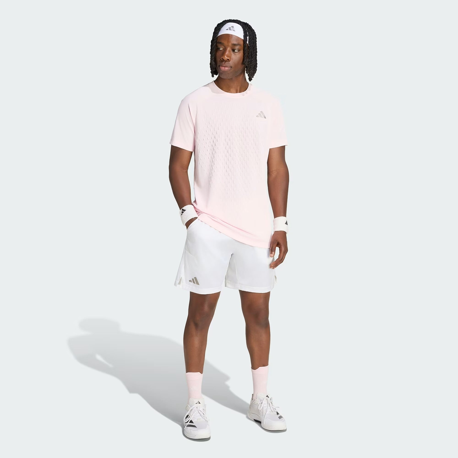 A man stands on a light gray background wearing a light pink T-shirt, adidas Climacool Ergo Pro Mens Badminton Shorts in white, white sneakers, pink socks, wristbands, and a white headband. He looks to the side with one hand in his pocket.