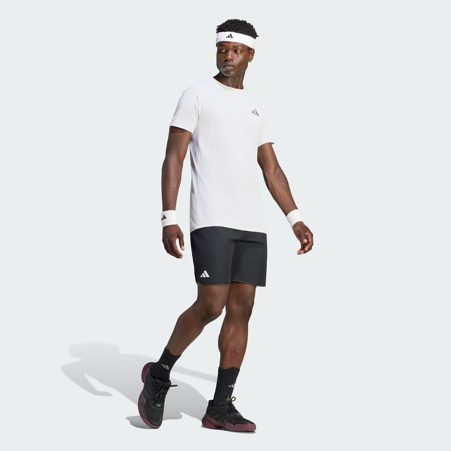 A man stands confidently against a plain light gray background, dressed in the ADIDAS Climacool+ Freelift Pro Men’s Badminton T-Shirt in white by adidas, paired with black shorts, black socks, and black sneakers. He accessorizes with a white headband and wristbands. The quick-drying sports tee ensures comfort during intense activity.