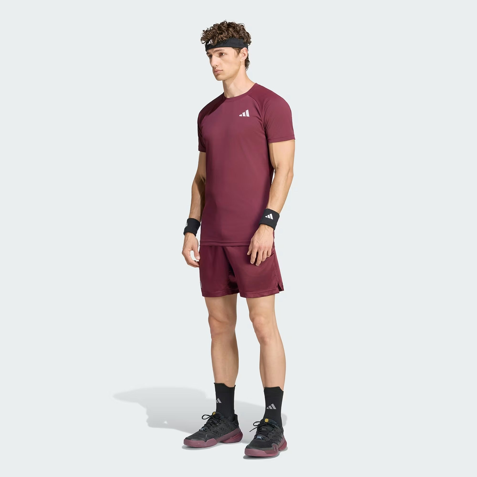 A man wearing the adidas ADIDAS Climacool+ Freelift Pro Men’s Badminton T-Shirt in maroon, paired with matching shorts, black wristbands, socks, sneakers, and a headband stands against a plain light background.