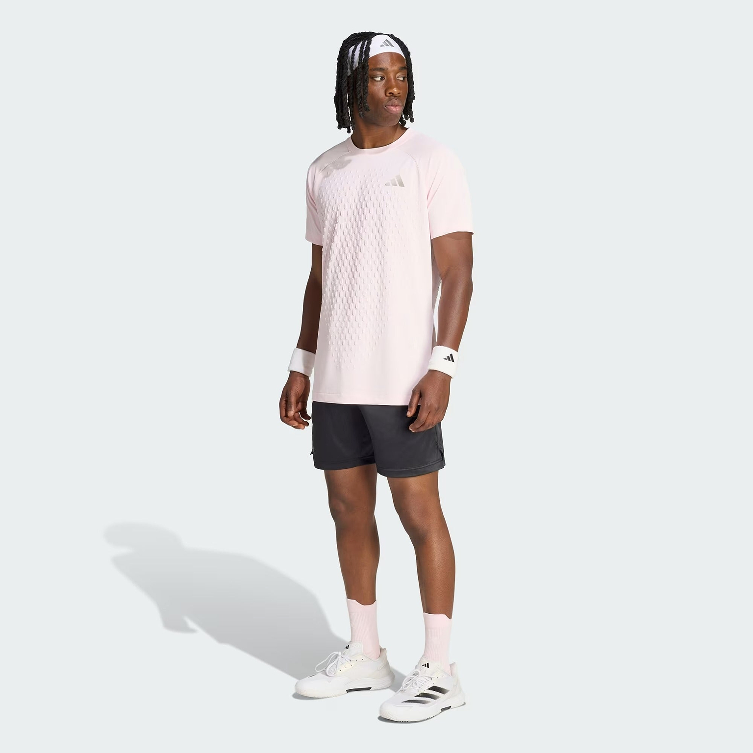 A man wearing a light pink Adidas shirt, black adidas Climacool Ergo Pro men’s badminton shorts, white wristbands, white socks, and white adidas sneakers stands facing slightly left against a plain light background.