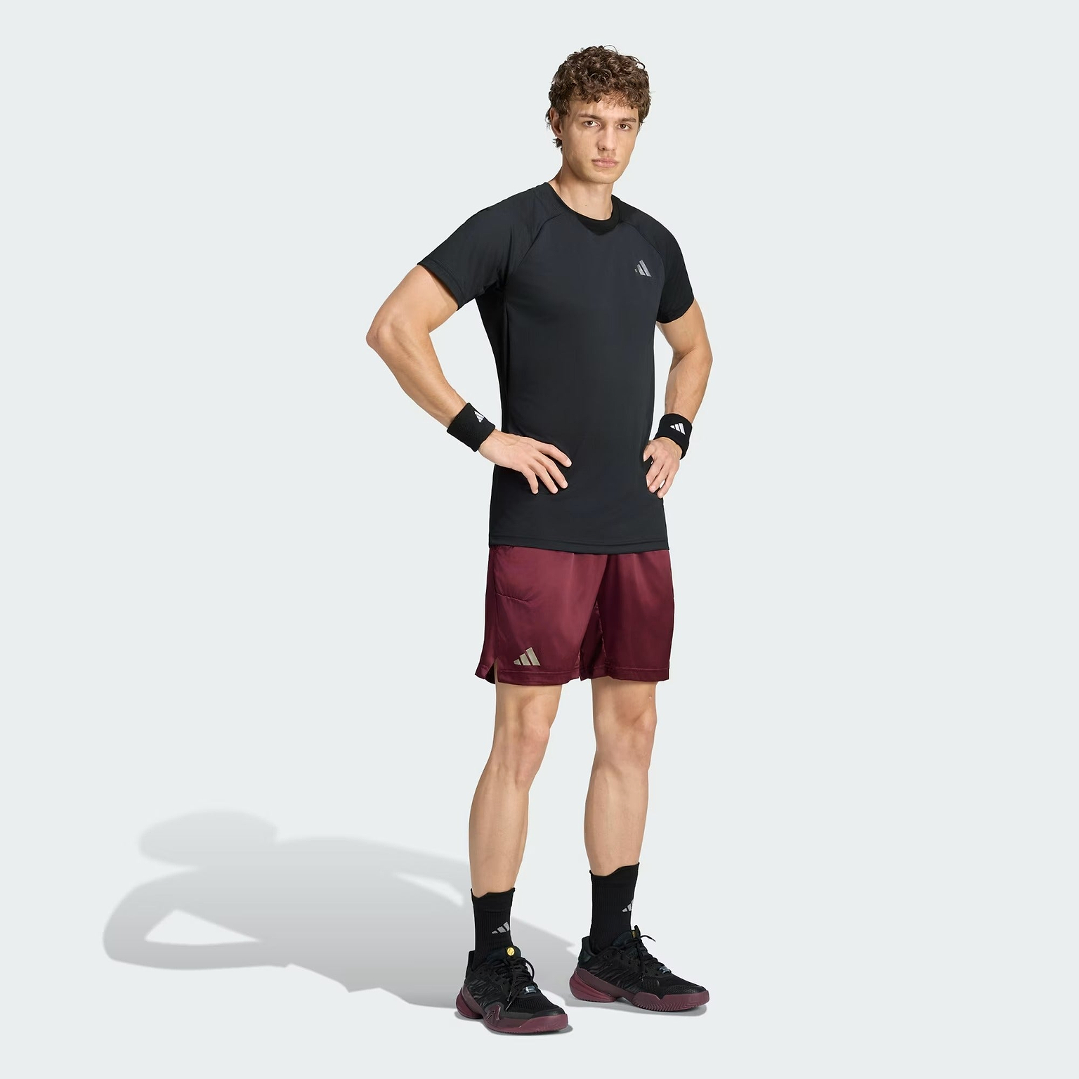 A person stands with hands on hips, wearing an adidas Climacool+ Freelift Pro Men’s Badminton T-Shirt in black, featuring advanced cooling technology, maroon shorts, black shoes, black wristbands, and black socks against a plain light background.