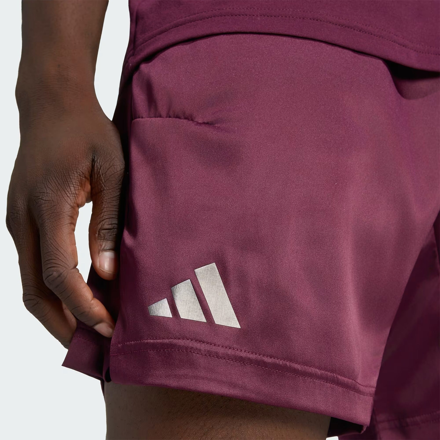 A person wearing ADIDAS Climacool Ergo Pro Mens Badminton Shorts in maroon, featuring a white adidas logo on the left leg, stands against a plain light background.