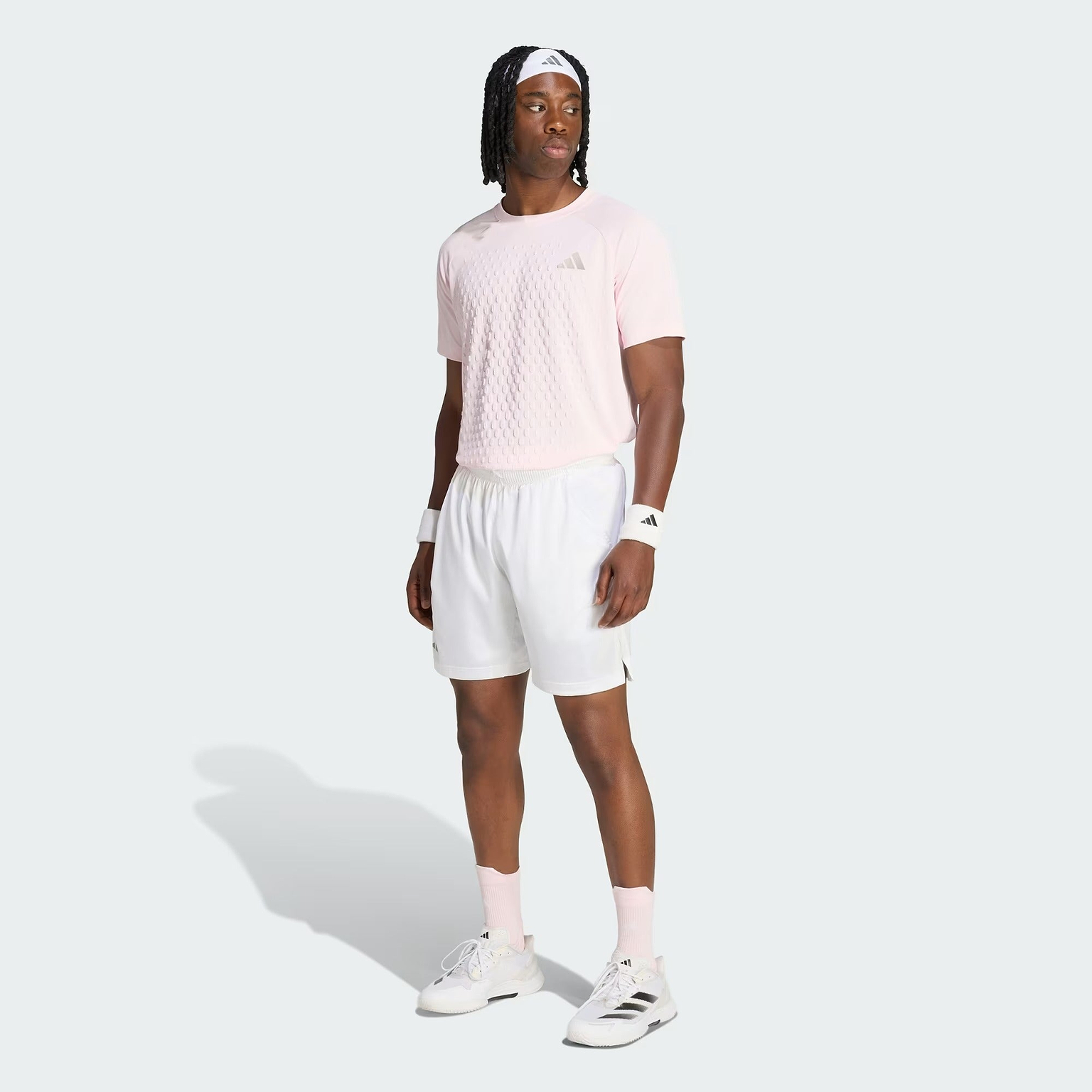 A man in a pink adidas shirt, adidas Climacool Ergo Pro Men’s Badminton Shorts in white, white sneakers, wristbands, and a headband stands on a light gray background, looking to the side.