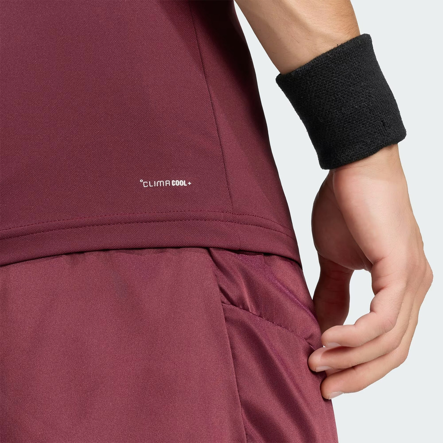 A person is shown from the side, wearing a maroon adidas Climacool+ Freelift Pro Mens Badminton T-Shirt and a black wristband, with their hand near their hip. The visible “Climacool” branding on the shirt emphasizes its identity as a high-performance badminton T-shirt from adidas.
