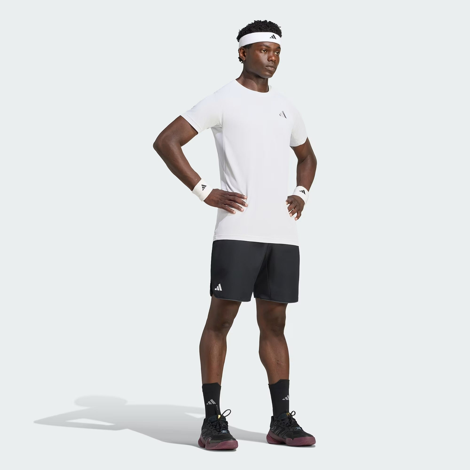 A man wearing an adidas Climacool+ Freelift Pro Men’s Badminton T-Shirt in white, black shorts, white headband, white wristbands, and black shoes stands with his hands on his hips against a plain background.