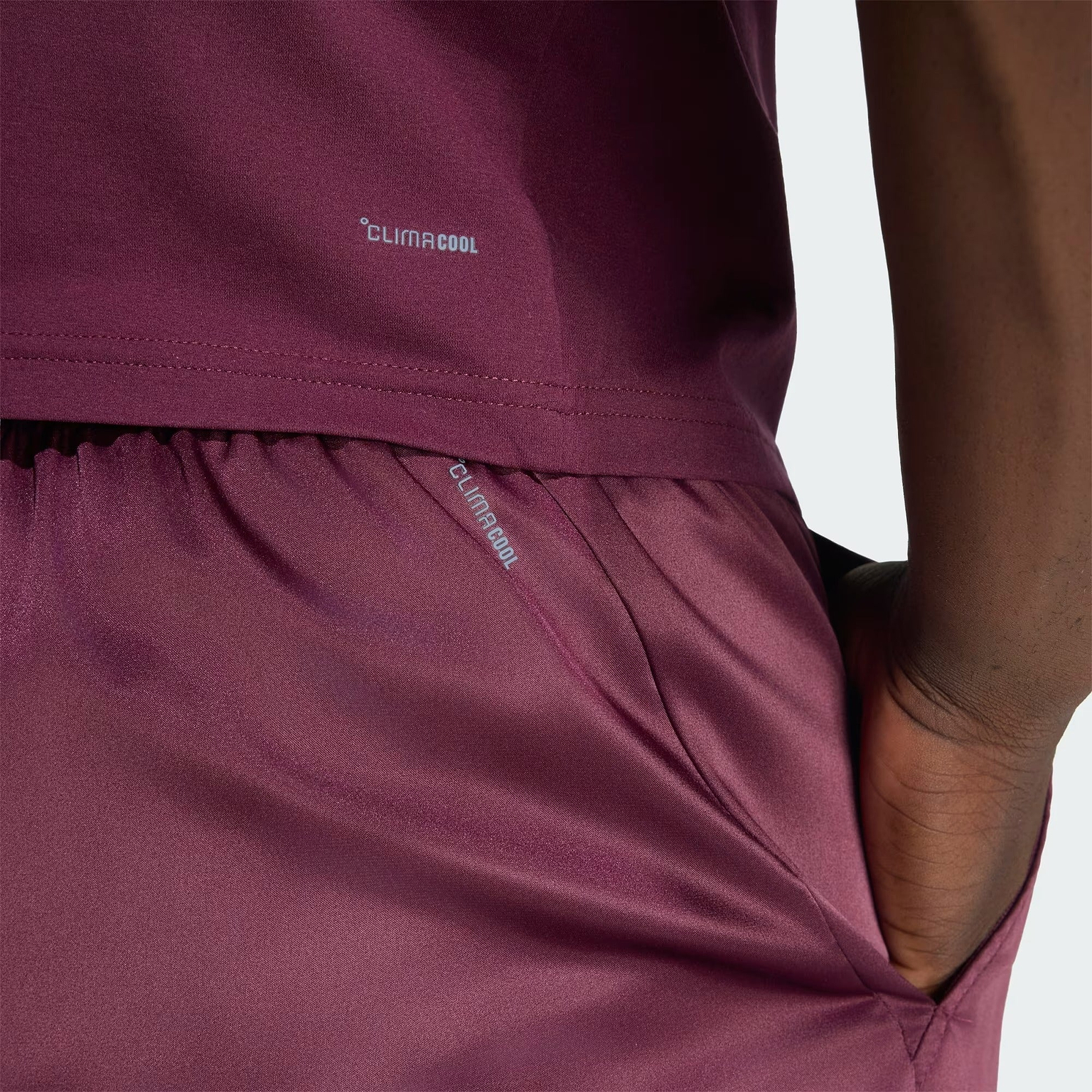 A close-up shows a person wearing maroon ADIDAS Climacool Ergo Pro Men’s Badminton Shorts, with their hand in the pocket. The word “Climacool” is visible on both the shirt and these ergonomic adidas badminton shorts. The background is plain and light-colored.