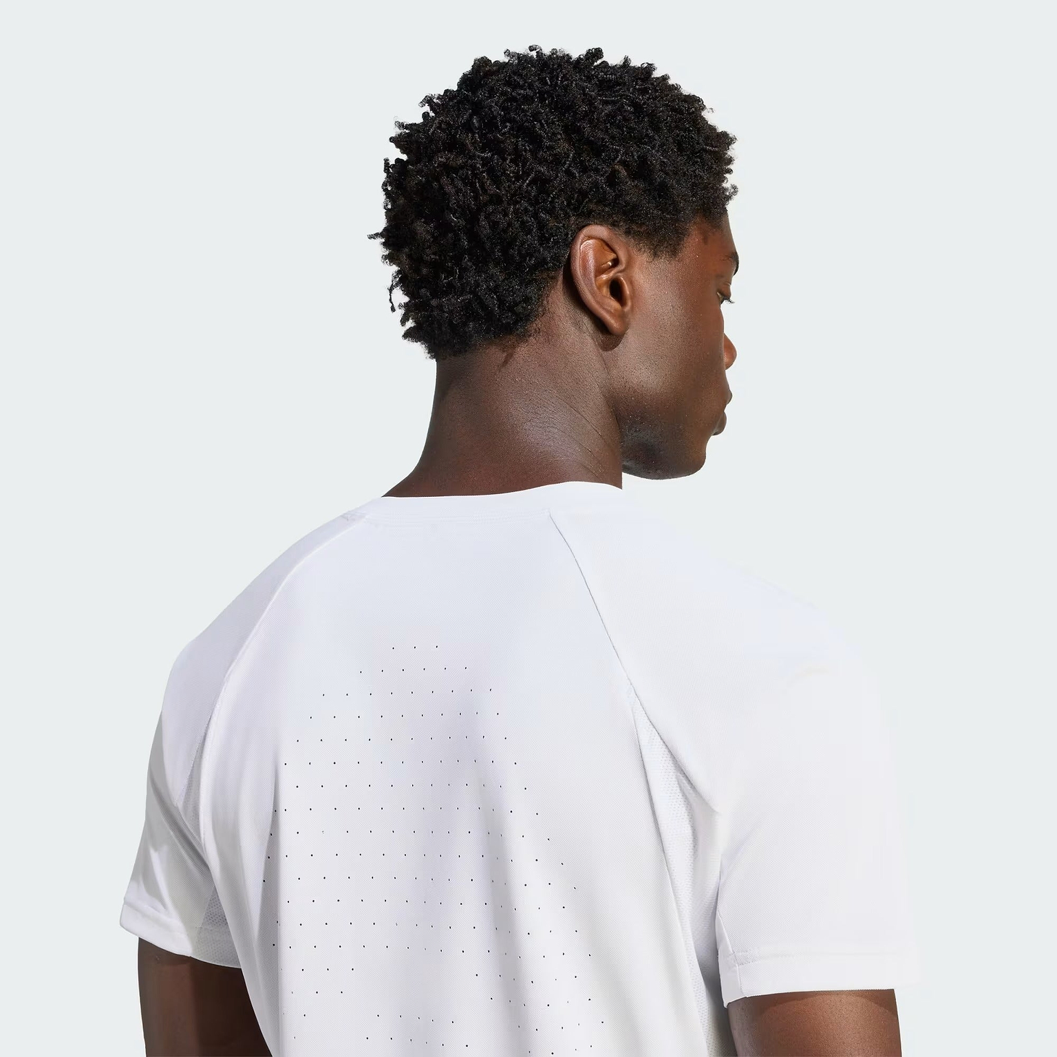A person with short curly hair is shown from behind, wearing the adidas ADIDAS Climacool+ Freelift Pro Men’s Badminton T-Shirt in white, featuring small perforations on the back for ventilation, against a plain light background.