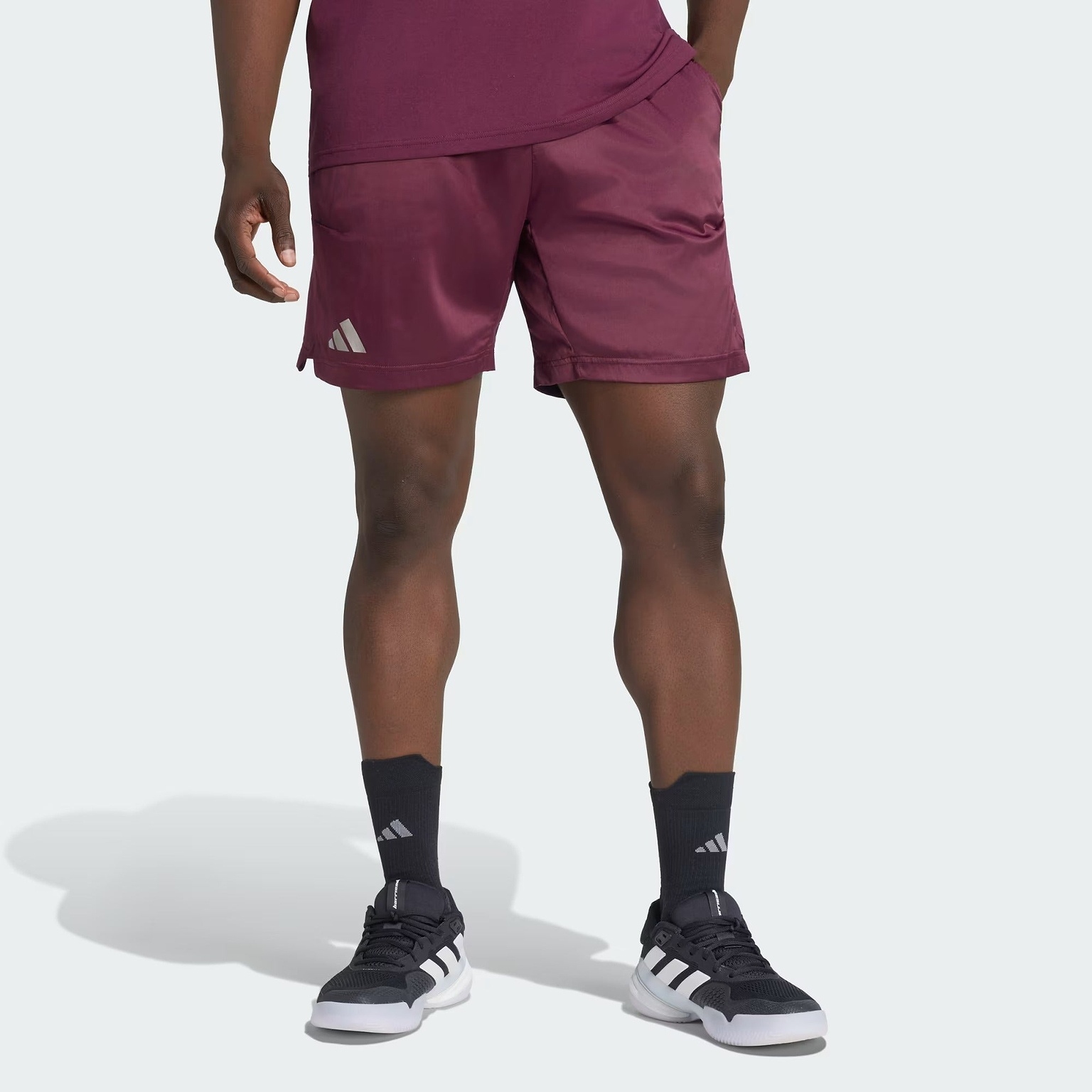 A person is wearing adidas Climacool Ergo Pro Men’s Badminton Shorts in maroon, featuring the adidas logo, paired with black adidas socks and black and white athletic sneakers. The ergonomic sports shorts utilize Climacool technology for enhanced breathability. The background is a solid light gray.