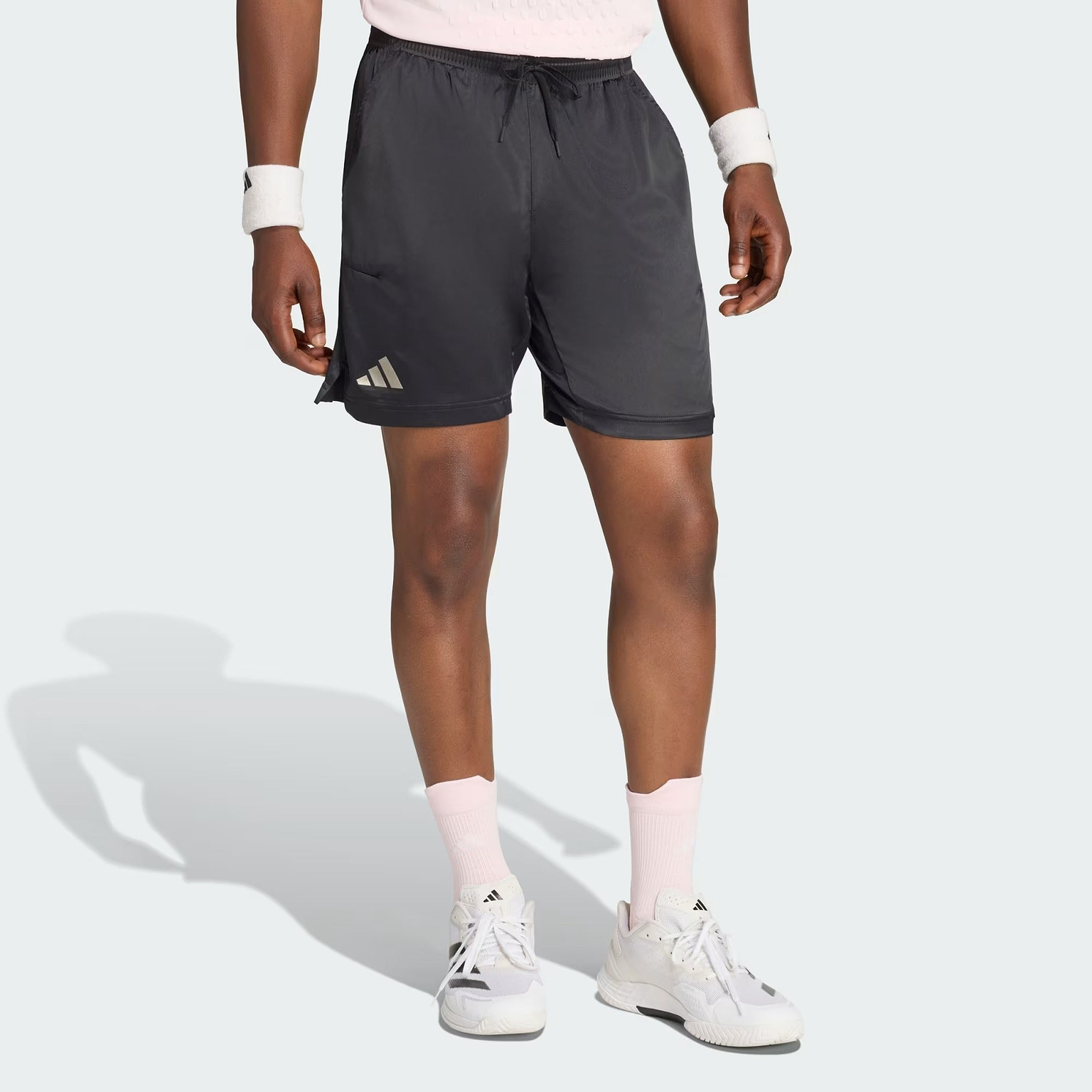 Person wearing adidas Climacool Ergo Pro Men’s Badminton Shorts in black, paired with white sneakers, white and pink socks, and white wristbands, standing on a light gray background with only the lower body visible.