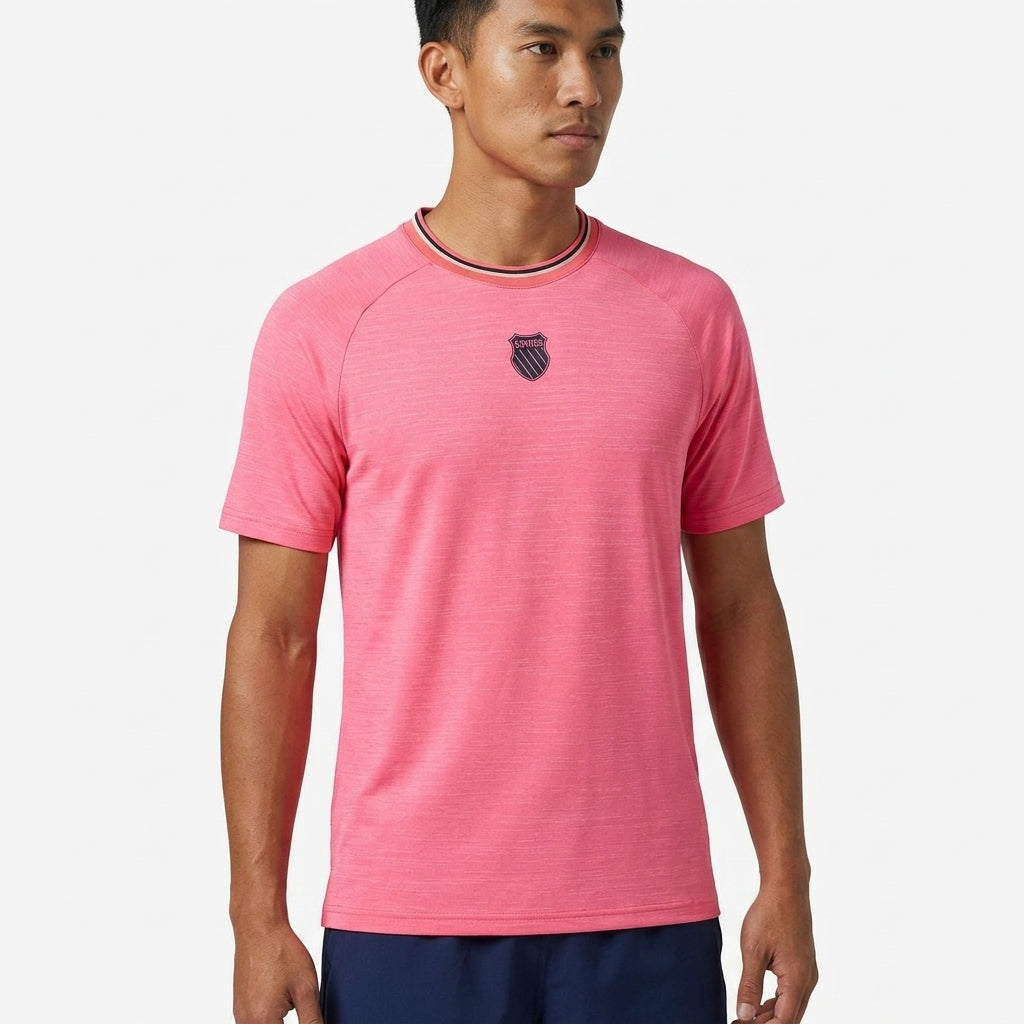 A man wears the K-Swiss Hypercourt Melange 3 Men's Badminton T-Shirt in pink, featuring a small dark chest emblem, paired with navy shorts and standing against a plain white background.